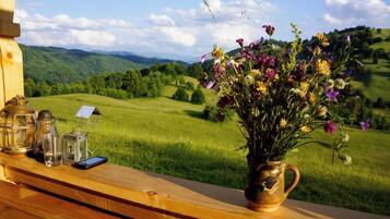 Une table en bois, ornée d’un vase de fleurs et de lanternes, se détache sur un paysage pittoresque composé d’un champ verdoyant et de collines.