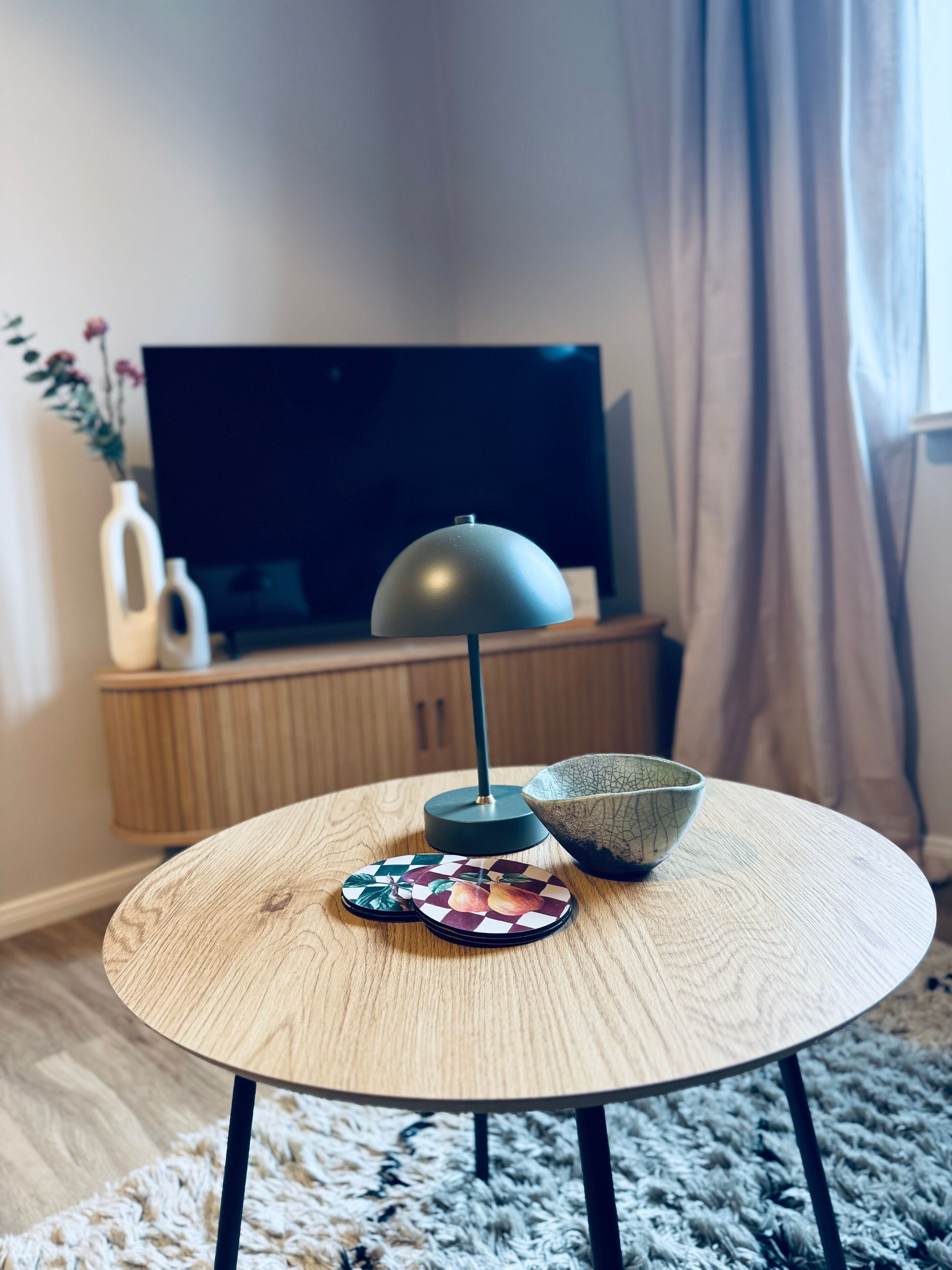 A round wooden table with a modern lamp, a decorative bowl, and a floral coaster set.