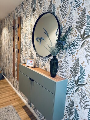 A room with a patterned wallpaper, a round mirror, a wooden cabinet, and a vase with plants.