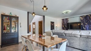 A modern living room with a large flat-screen TV, a grey sofa, and a wooden dining table with chairs.