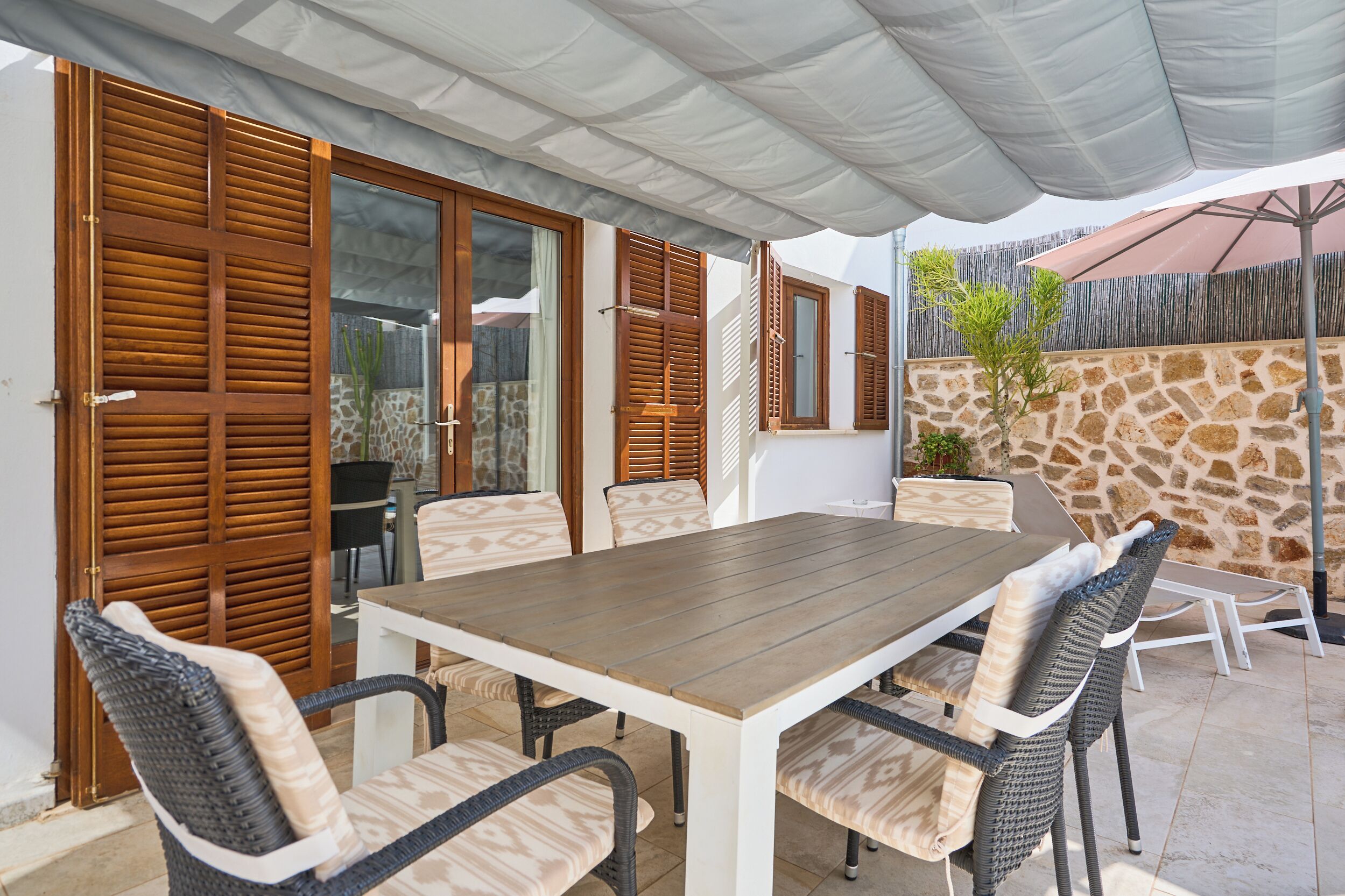 A patio with a wooden table and wicker chairs, a stone wall, and a covered area with umbrellas.