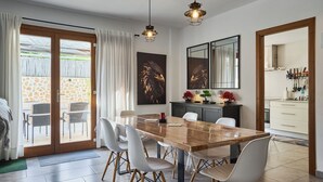 A dining area with a wooden table and white chairs, a kitchen with white cabinets, and a stone wall visible through the glass door.
