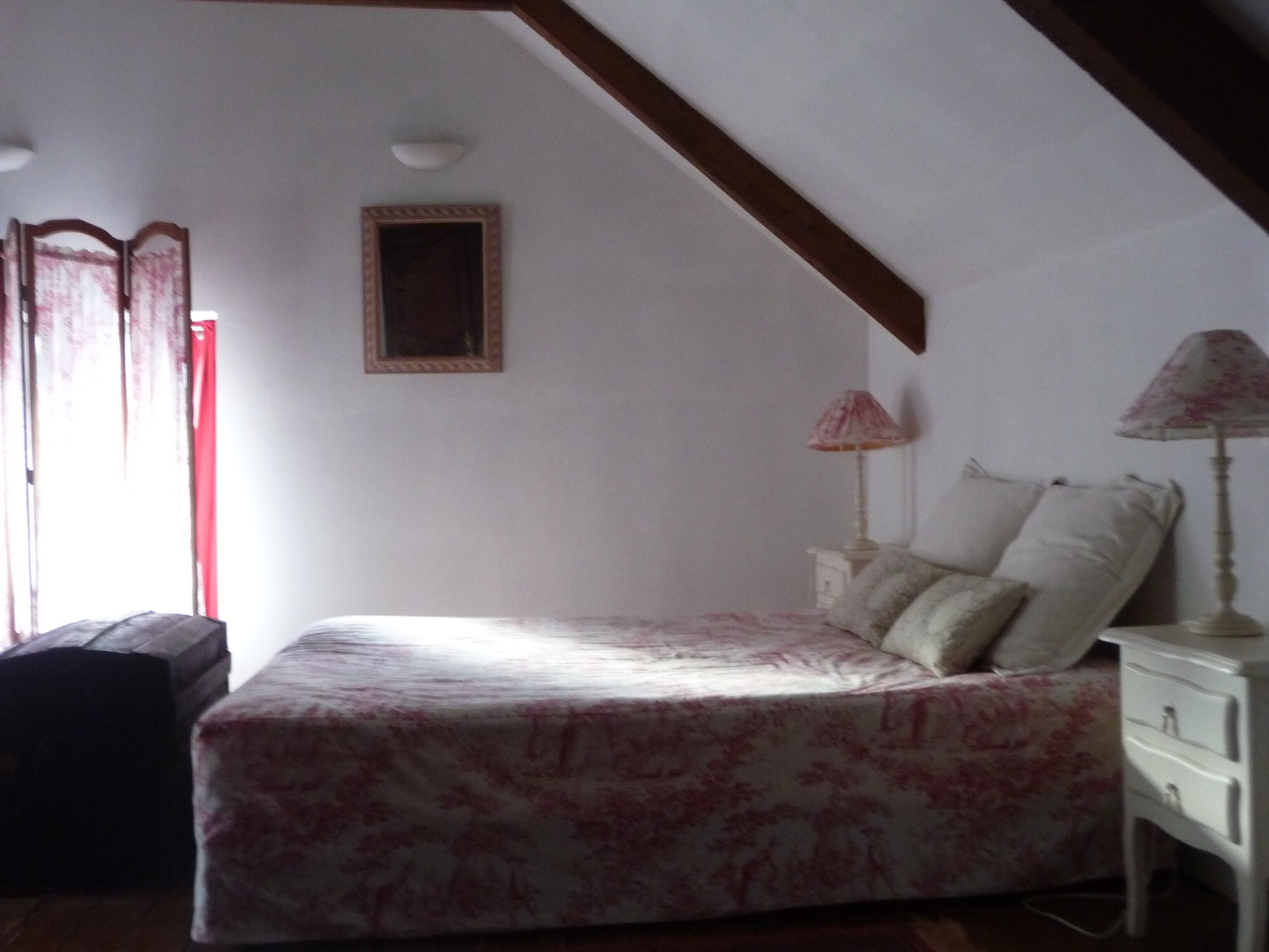 Un dormitorio con cama, mesita de noche, lámpara, ventana con cortinas y una viga de techo de madera.