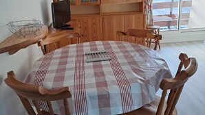 Ruokailutilassa on pyöreä pöytä, jonka päällä on ruutukuviollinen pöytäliina, ja pöydän ympärillä on puisia tuoleja. Huoneessa on myös puinen kaappi, jossa on lasiovet ja jonka sisällä on astioita ja laseja. Sivupöydällä on sininen lamppu ja seinällä kello.