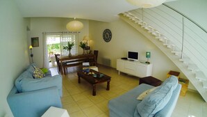 Una sala de estar con un sofá, una mesa de centro y un televisor sobre un mueble blanco. Se observa una escalera y un comedor con una mesa y sillas.