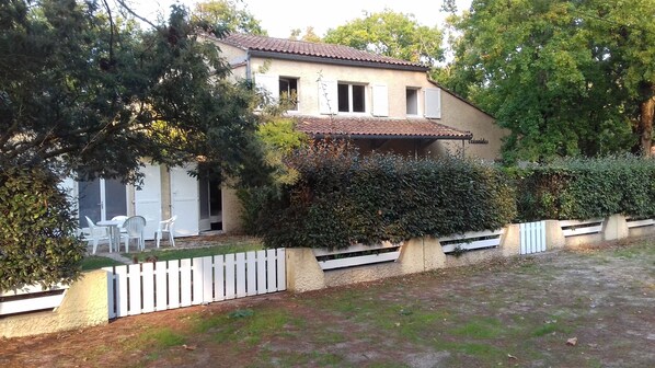 Una casa a due piani con tetto in tegole, persiane bianche e un piccolo balcone. All'esterno, ci sono sedie e un tavolo in plastica bianca, e un recinto di legno bianco delimita la proprietà.