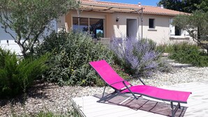 A house with a tiled roof, a covered porch, and a garden with various plants and a pink lounge chair.