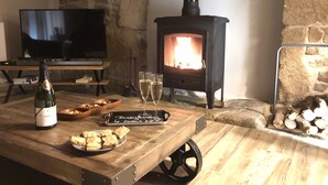 Une pièce chaleureuse avec un mur en pierre, une table en bois sur laquelle se trouvent une bouteille de vin, des verres et des amuse-gueules, ainsi qu'un poêle à bois noir.