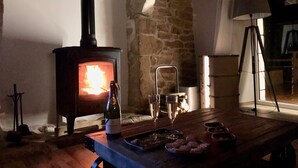 Une pièce chaleureuse avec un mur en pierre, un poêle à bois, une table sur laquelle se trouvent une bouteille de vin, des verres et des amuse-gueules.