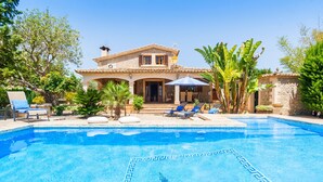 A swimming pool with a tiled pattern, a house with a tiled roof, and a patio area with lounge chairs and umbrellas.