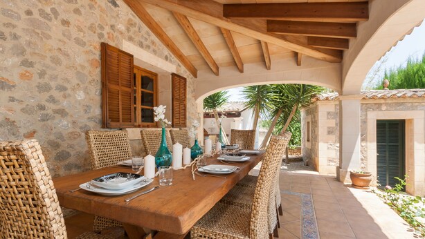 A dining area with a stone wall, wooden ceiling beams, wicker chairs, and a long wooden table set for six.