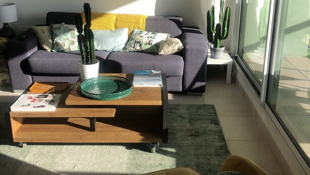 Ein modernes Wohnzimmer mit einer Couch, einem Couchtisch und einem gelben Sessel. Auf dem Couchtisch steht ein Kaktus und auf einem Beistelltisch ein weiterer. Ein Bild mit Kakteen hängt an der Wand.