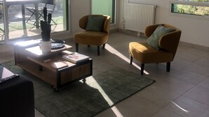 Ein modernes Wohnzimmer mit Sofa, Sessel und Couchtisch. Ein Fernseher ist an der Wand montiert und ein Fenster bietet Blick auf Gebäude.
