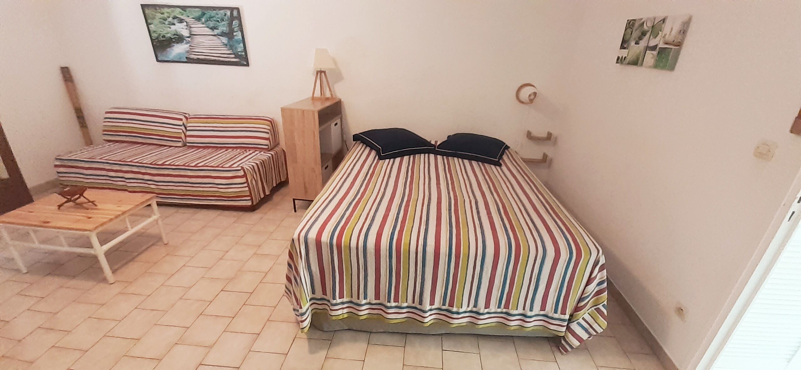 A small, cozy room with a striped bed, a sofa, a wooden table, and a wall-mounted lamp.