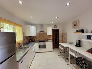 A modern kitchen with a refrigerator, washing machine, stove, and sink. There is a clock on the wall and a shelf with kitchen items.