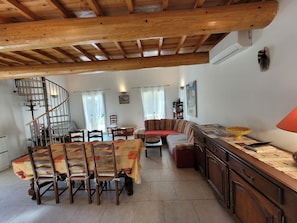 A spacious living area with a wooden ceiling, a dining table with chairs, a sofa, a lamp, and a staircase leading to an upper level.