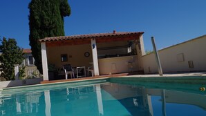 Ein Poolbereich mit überdachter Terrasse, einem hohen Baum und einem Gebäude mit einem Ziegeldach.