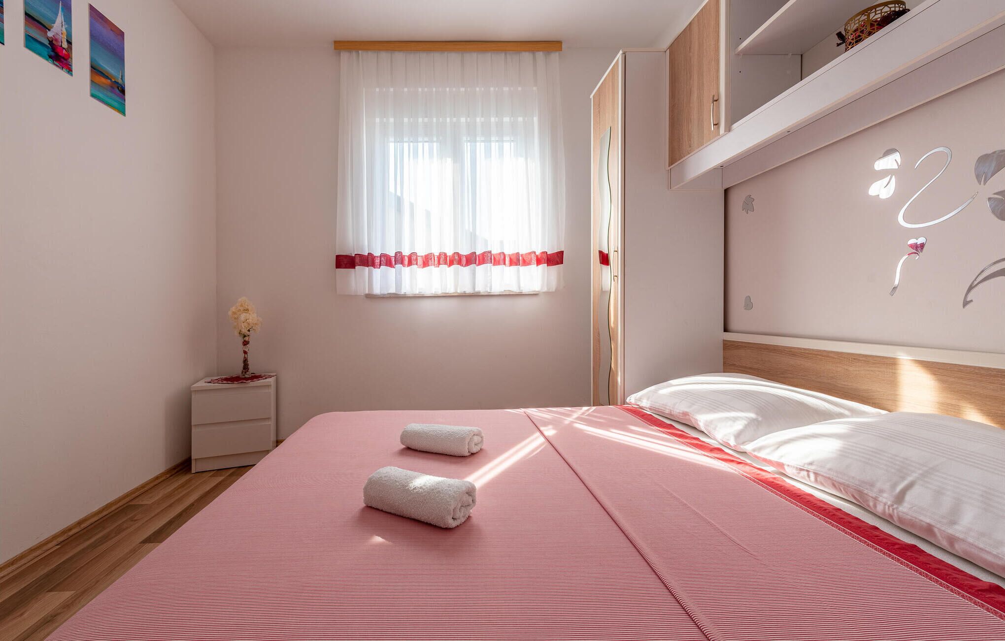 Een slaapkamer met een bed, een nachtkastje, een raam met dunne gordijnen en een wand met decoratieve details.