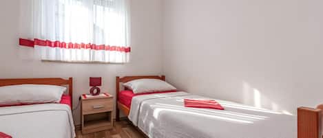 Ein Schlafzimmer mit zwei Betten, einem Nachttisch mit Lampe, einem Fenster mit durchsichtigen Vorhängen und einem roten Tagesdecke, sowie einem kleinen Gemälde an der Wand.