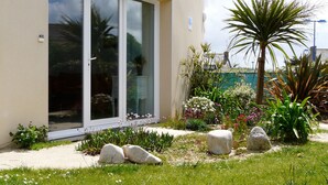 Un edifici modern amb grans portes de vidre, un jardí ben cuidat amb diverses plantes i roques, i un cel serè.