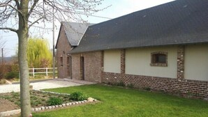 Un bâtiment d’un seul étage, construit en briques, avec un toit en pente, un petit jardin agrémenté d’un arbre et d’un parterre de fleurs, et une clôture blanche en arrière-plan.