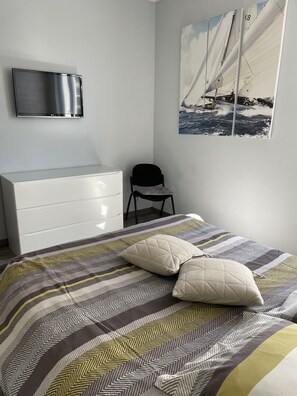 A bedroom with a bed, a TV mounted on the wall, a chair, and a painting of a sailboat.