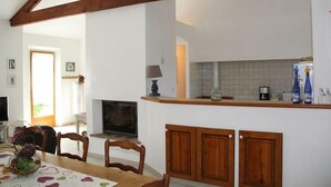 Zona pranzo con tavolo in legno, sedie e televisore. Si intravede un angolo cottura con pensili e rivestimento in piastrelle.