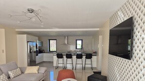 Ein modernes Wohnzimmer mit einer Couch, einem Hocker und einem Couchtisch. Im Hintergrund ist eine Küche mit einer Bar und Stühlen zu sehen.