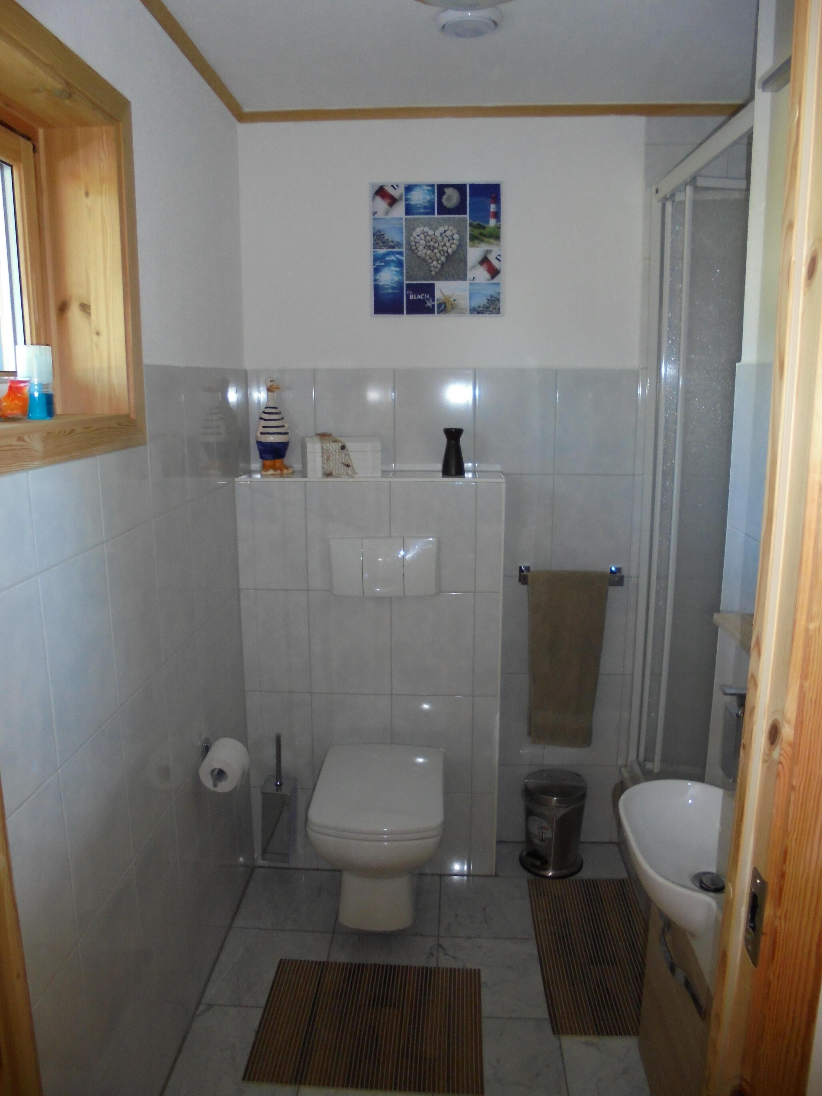 A small bathroom with a toilet, sink, and shower. There is a towel hanging on the wall and a small shelf with toiletries.
