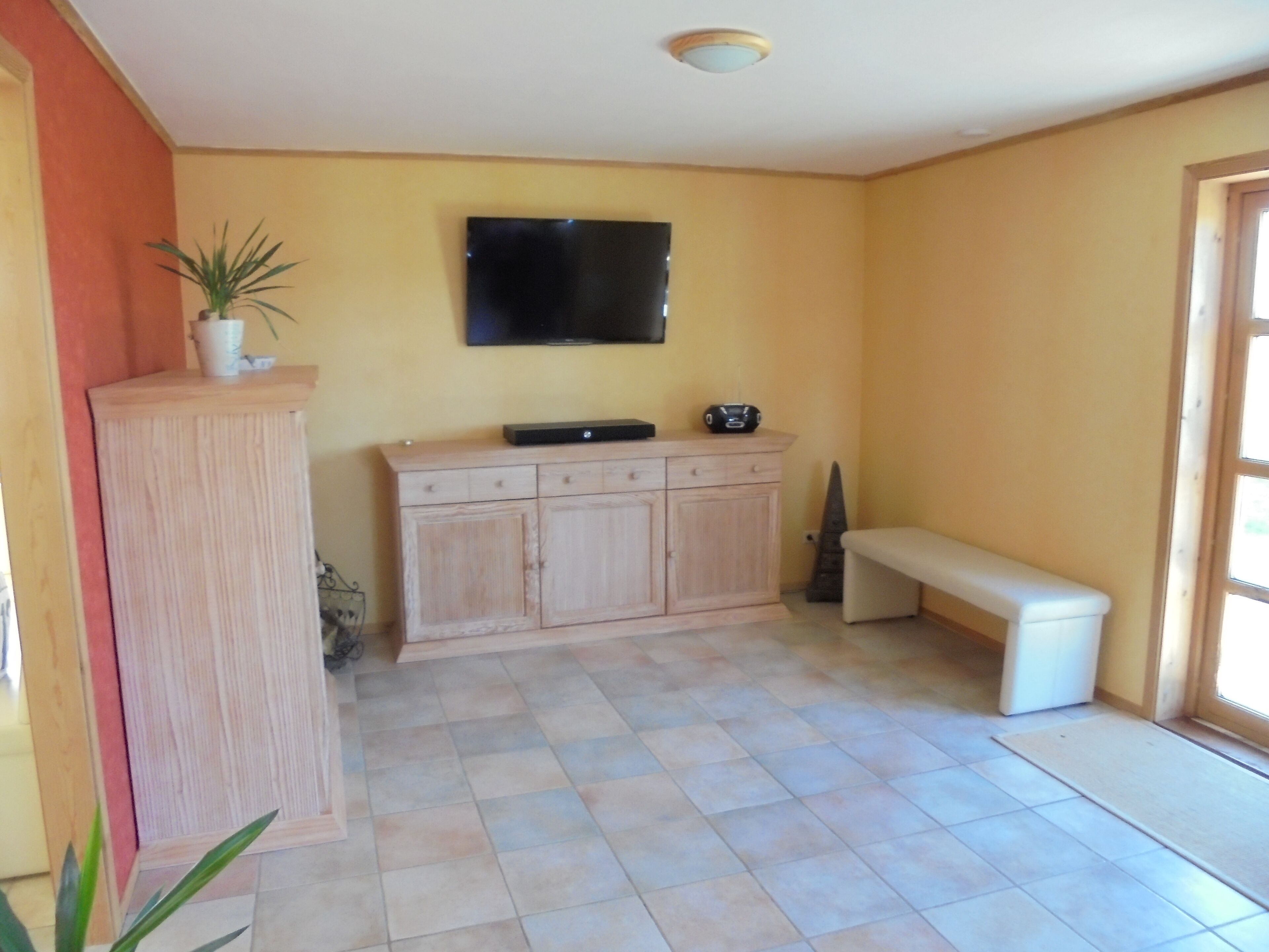Een kamer met een betegelde vloer, een houten kast, een bankje, een kamerplant en een flatscreen televisie die aan de muur is bevestigd.