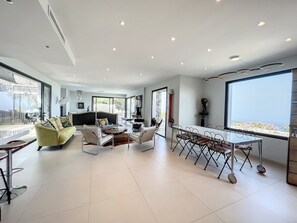 A modern living room with a sofa, chairs, a coffee table, and a dining area with a table and chairs. There is a large window providing a view of the outdoors.