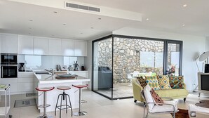 A modern kitchen with white cabinets, a central island with stools, and a stone wall adjacent to a sliding glass door leading to an outdoor seating area. - Luxury villa, sea view - swimming pool facing the sunset (Corbara)