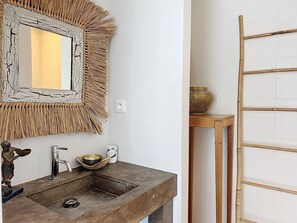 Une salle de bain avec un lavabo en pierre, un miroir doté d’un cadre texturé, une petite statue et une étagère en bois sur laquelle se trouve un bol.