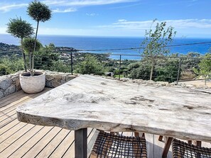 Une terrasse en bois avec une table en pierre et des plantes en pot, offrant une vue sur un paysage côtier.