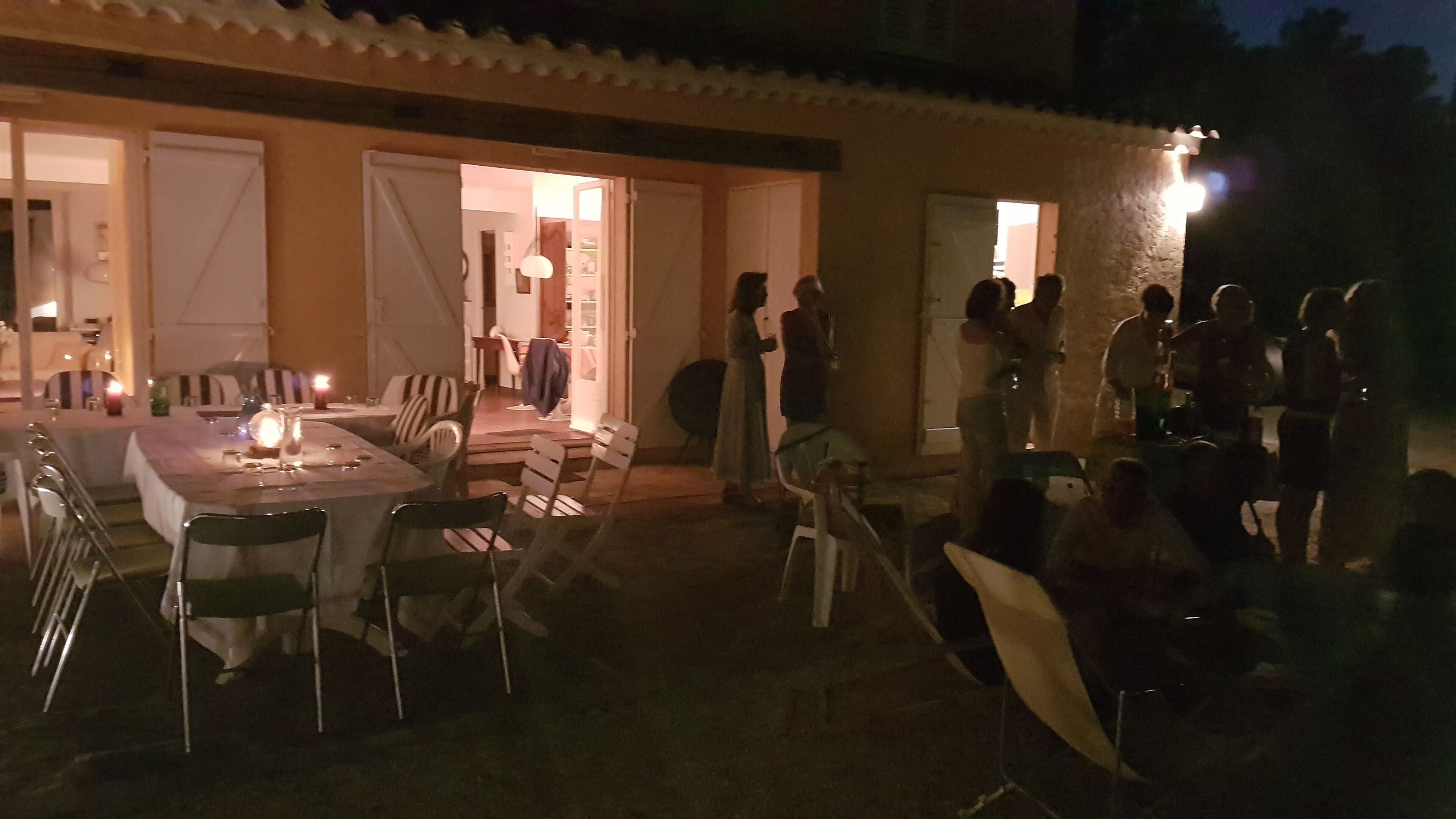 An outdoor dining area with tables and chairs, a few people standing and conversing, and a building entrance in the background.