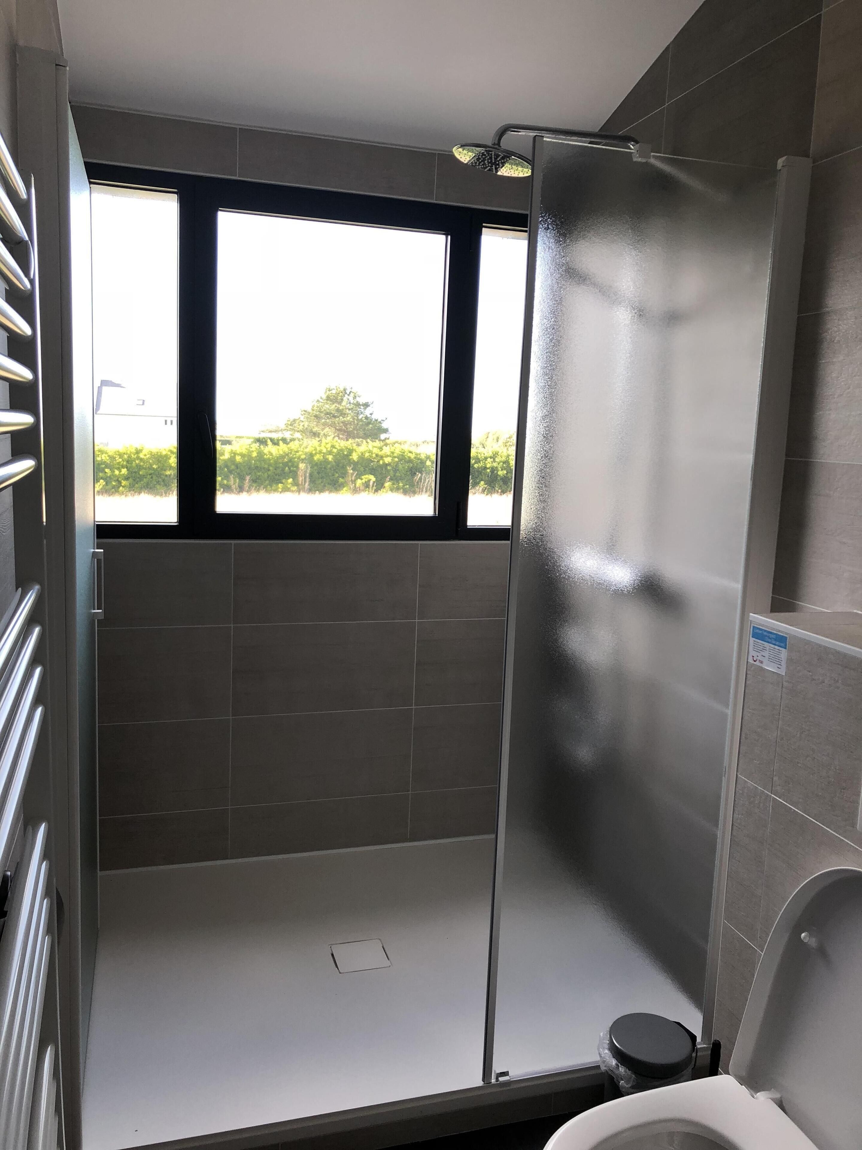 A modern bathroom with a shower, toilet, and a window offering a view of greenery.