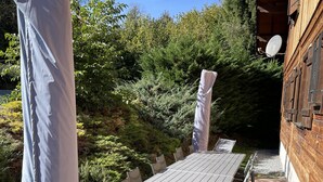 An outdoor dining area with a long table and chairs, surrounded by greenery and a wooden building.