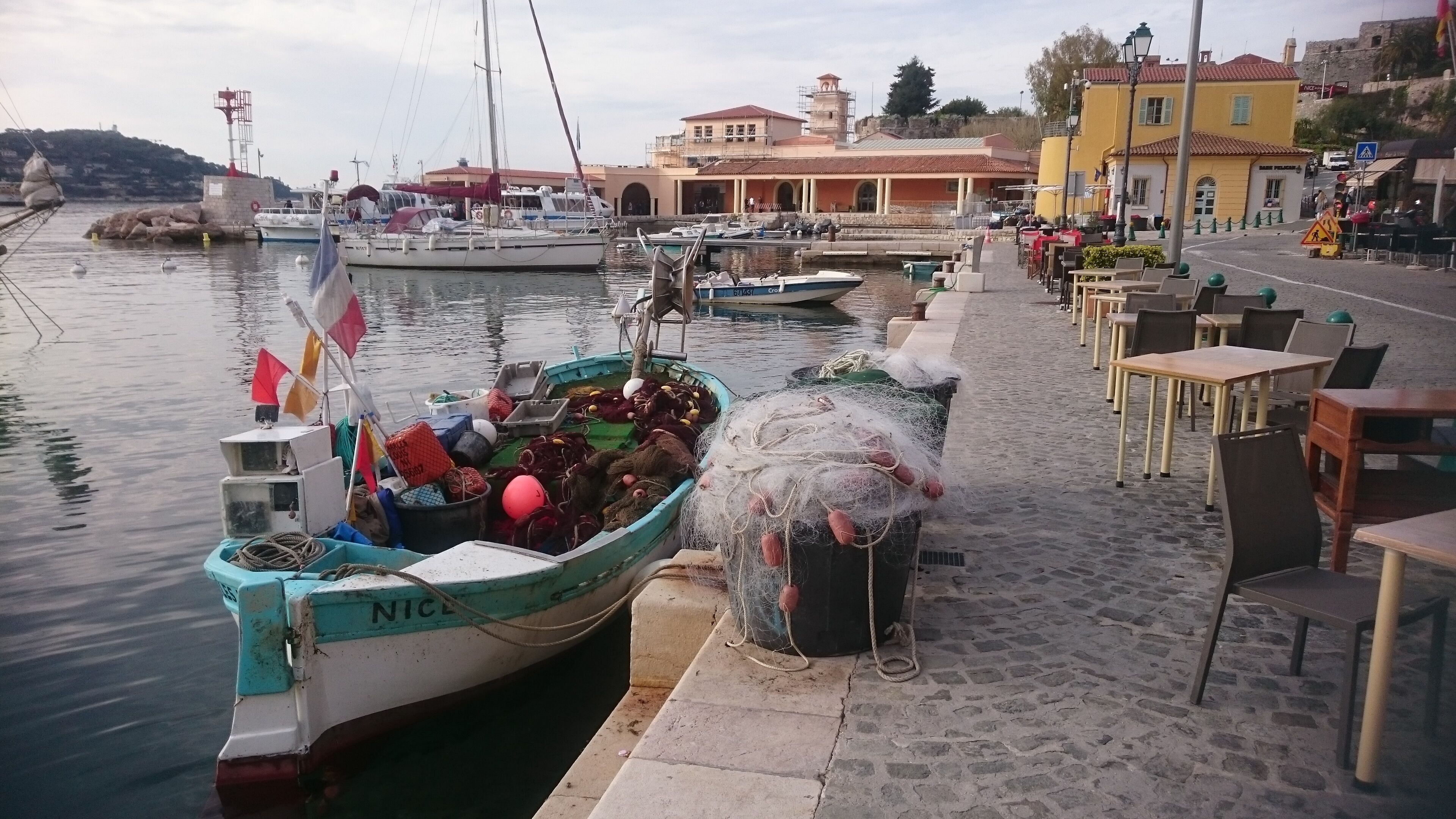 A harbor with boats docked, a building with a red roof, and outdoor seating along the waterfront.