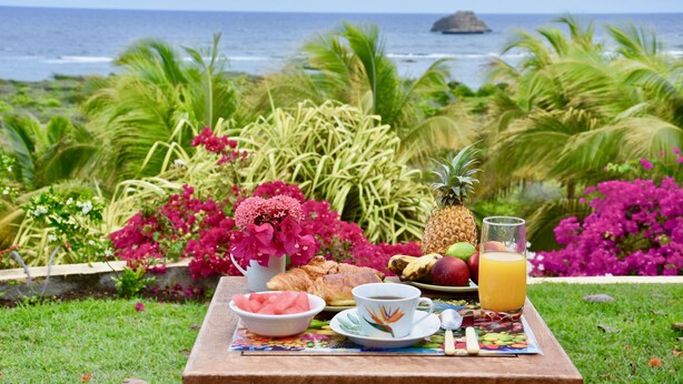 A breakfast setup with tropical fruits, a croissant, and a glass of orange juice on a table with a view of the ocean and palm trees.