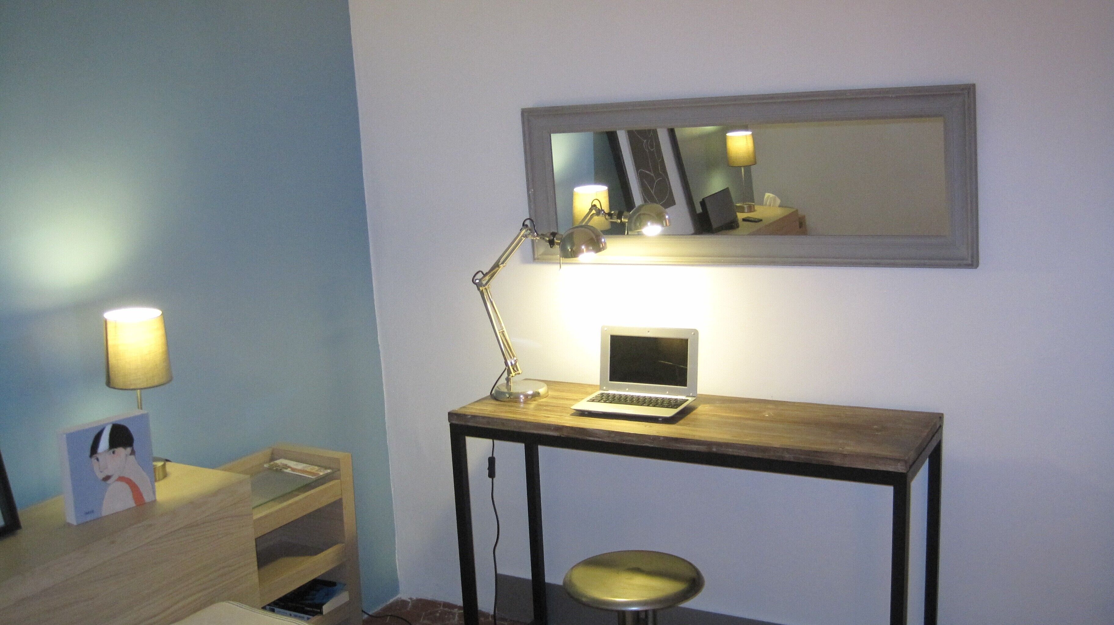 A small desk with a laptop, a lamp, and a mirror on the wall.