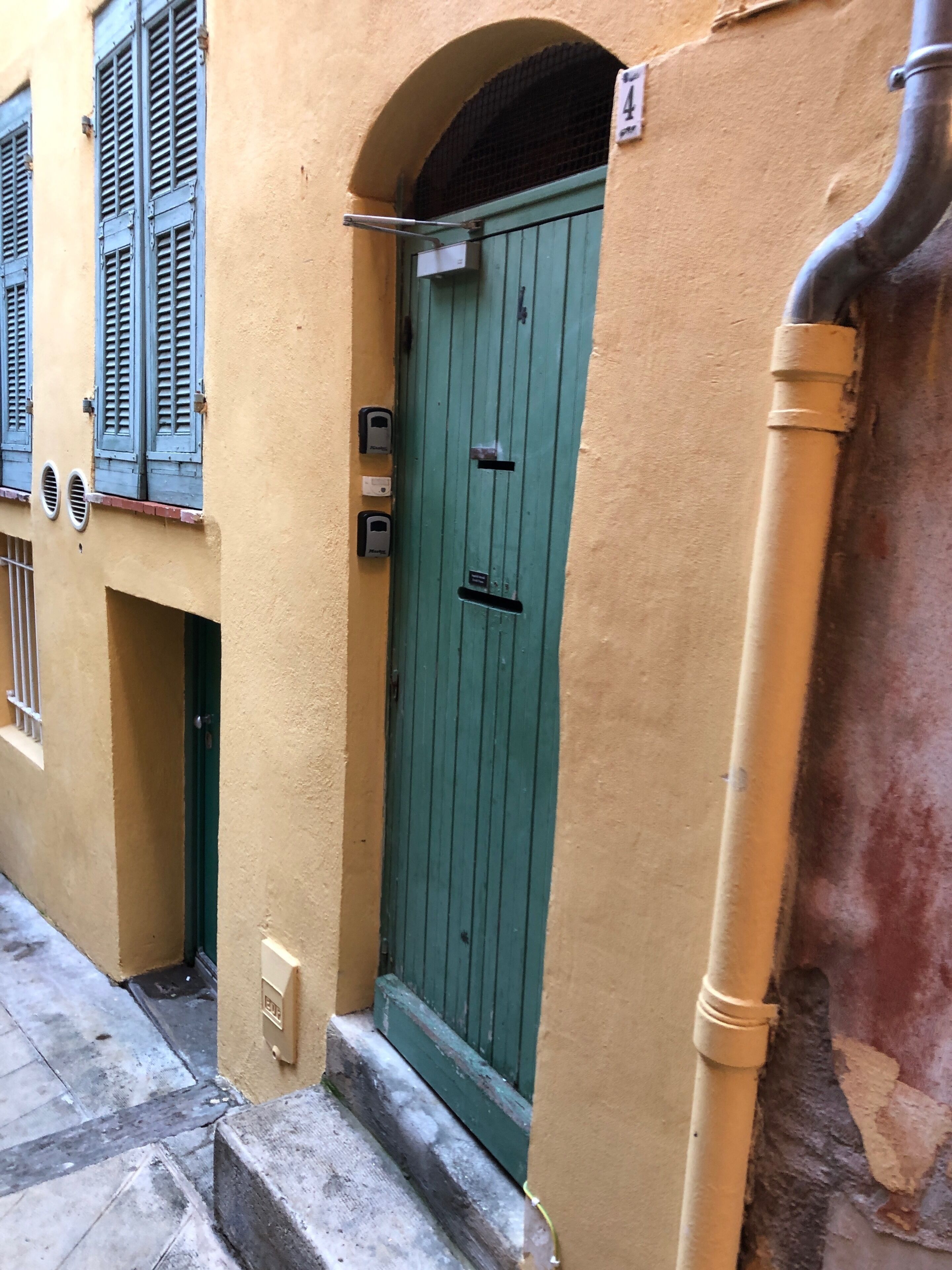 A narrow street with a green door, a yellow wall, and a metal pipe.