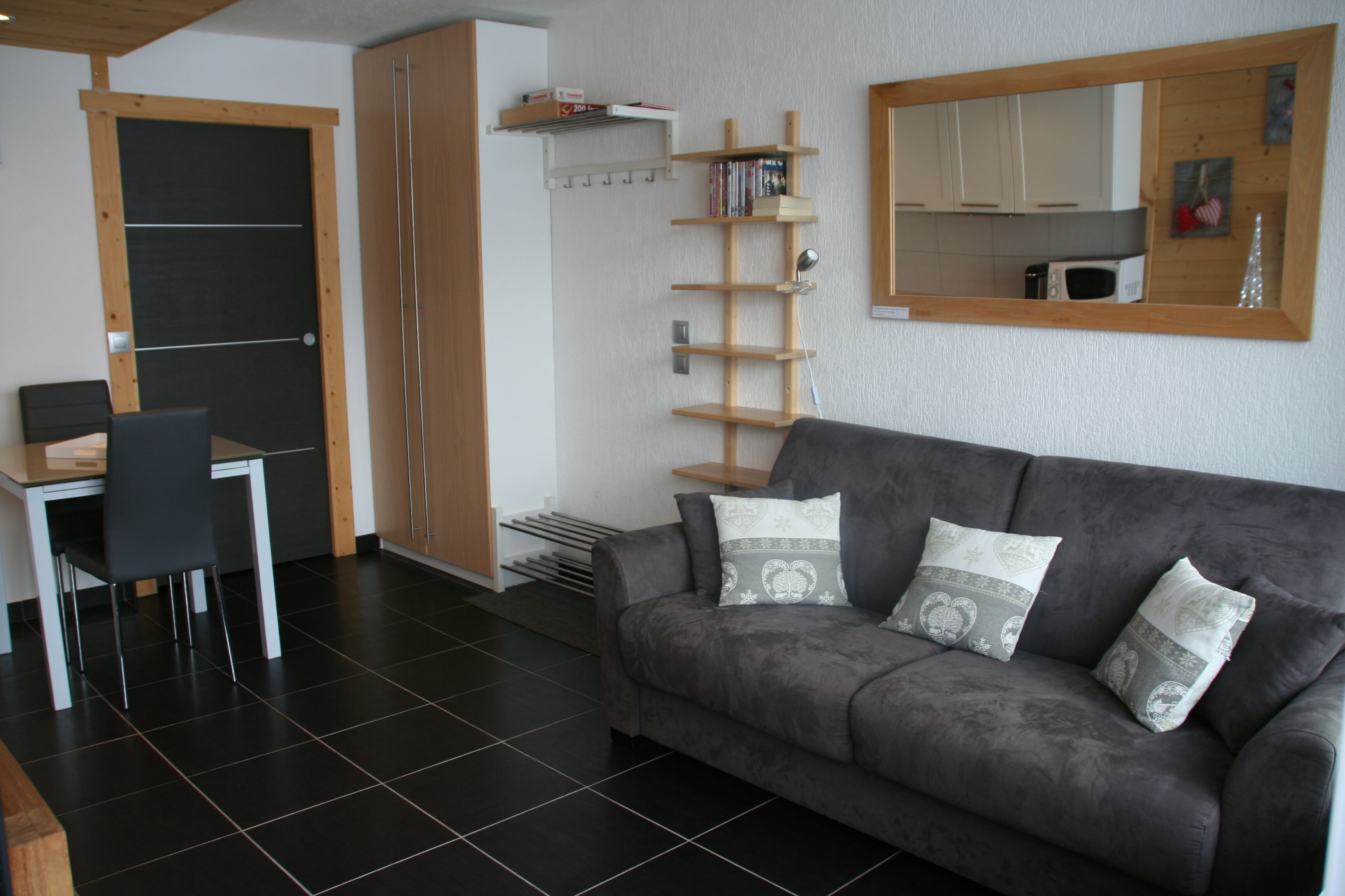 A modern living room with a grey sofa, two matching cushions, a wooden side table, a black chair, a built-in bookshelf, and a kitchen area with cabinets and a microwave.