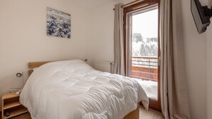 A bedroom with a wooden bed, a window with curtains, a radiator, and a wall-mounted TV. - Modern apartment in Plagne Centre with balcony, 2 bedrooms and Wifi (La Plagne)