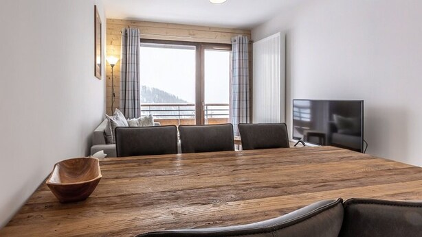 A modern room with a wooden dining table, four chairs, a television, and a large window with curtains. - Modern apartment in Plagne Centre with balcony, 2 bedrooms and Wifi (La Plagne)