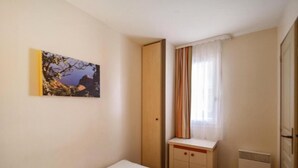 A bedroom with a bed, a small bedside table, a radiator, a window with curtains, and a painting on the wall. - Cap Esterel (St Raphaël) - Quiet apartment with sea view, golf & Cap Esterel 4 pers. (Saint-Raphaël)
