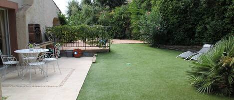 A patio area with a table and chairs, a small garden with artificial grass, and a variety of plants and trees.