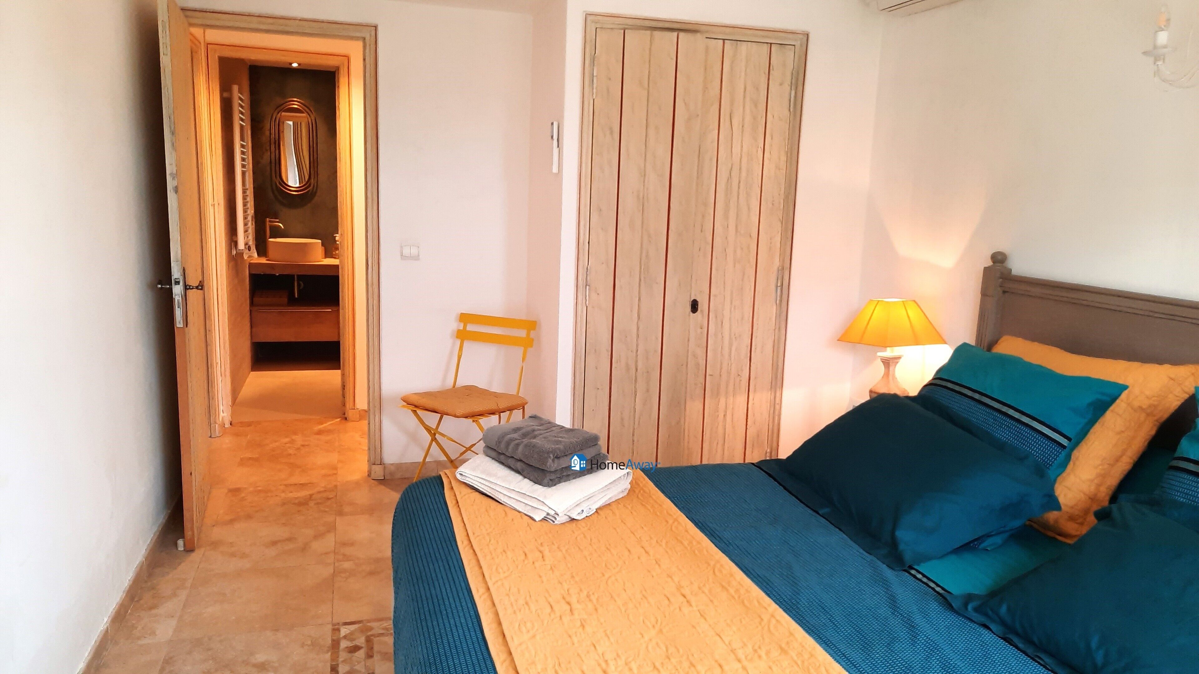 Ein Schlafzimmer mit einem Bett, einem Sessel, einem Tisch mit gefalteten Handtüchern, einem Holzschrank und einem durchgegangenen Tür sichtbar erkennbaren Badezimmer.