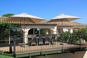 A building with a tiled roof, outdoor seating area with metal chairs and tables, and two large umbrellas providing shade.