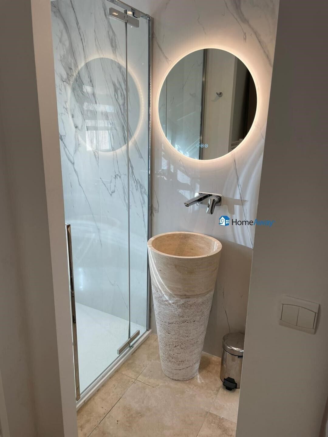 Un bagno con cabina doccia in marmo, un lavabo spazioso con specchio tondo e un cestino di piccole dimensioni.