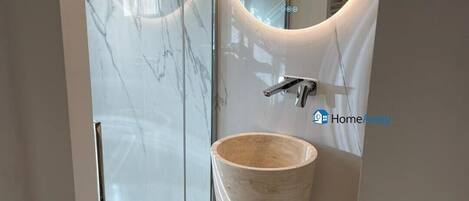 Un bagno con cabina doccia in marmo, un lavabo spazioso con specchio tondo e un cestino di piccole dimensioni.